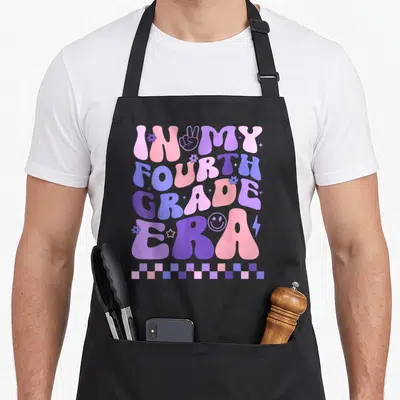 Era teacher apron - Bubbly IN MY FOURTH GRADE ERA text in pink purple blue with flowers stars peace sign — shown on a black cotton canvas apron against a white background