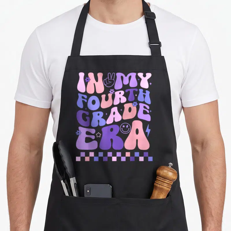 Era teacher apron - Bubbly IN MY FOURTH GRADE ERA text in pink purple blue with flowers stars peace sign — shown on a black cotton canvas apron against a white background