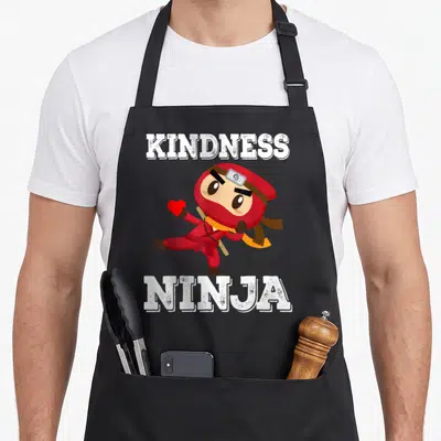 Bullying teacher apron - Cartoon ninja in red holding heart with white KINDNESS NINJA text — shown on a black cotton canvas apron against a white background