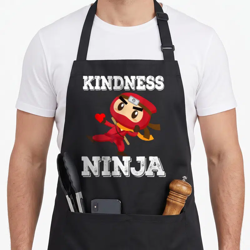 Bullying teacher apron - Cartoon ninja in red holding heart with white KINDNESS NINJA text — shown on a black cotton canvas apron against a white background