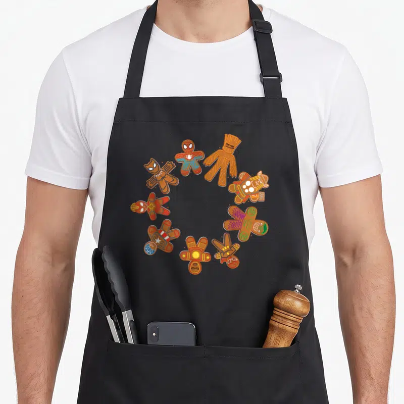 Circle gingerbread apron - Seven gingerbread superhero cookies in a ring with red blue and green icing — shown on a black cotton canvas apron against a white background