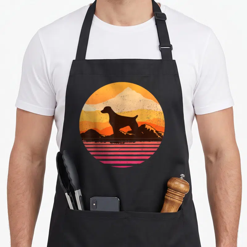 Sun dog apron - Retro circular sunset with black dog silhouette, orange layers, and pink horizontal stripes — shown on a black cotton canvas apron against a white background