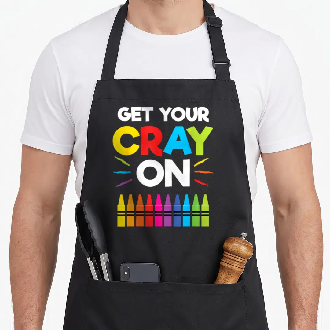 100th crayons apron - GET YOUR CRAY ON text with ten colorful crayons and scribbles — shown on a black cotton canvas apron against a white background