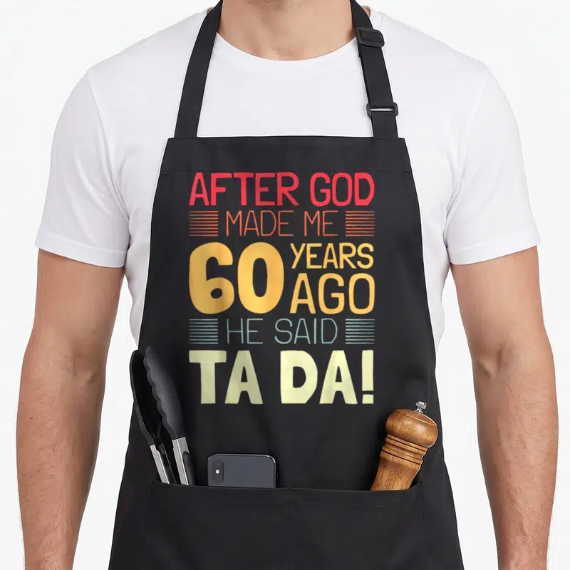 God 60th birthday apron - Stacked red orange yellow and blue text reading After God Made Me 60 Years Ago He Said Ta Da — shown on a black cotton canvas apron against a white background