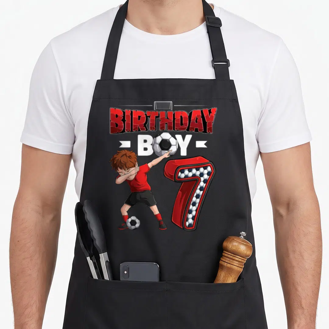 Dabbing soccer birthday apron - Cartoon boy dabbing next to number 7 and BIRTHDAY BOY text with soccer balls — shown on a black cotton canvas apron against a white background
