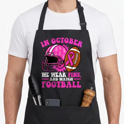 October football apron - Pink helmet with ribbons and football over pink splatter with awareness text — shown on a black cotton canvas apron against a white background