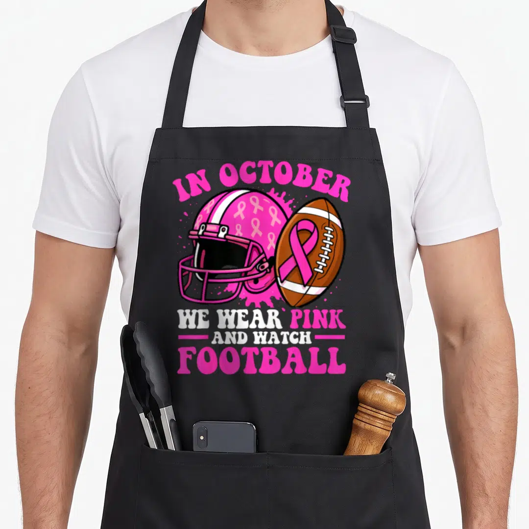 October football apron - Pink helmet with ribbons and football over pink splatter with awareness text — shown on a black cotton canvas apron against a white background