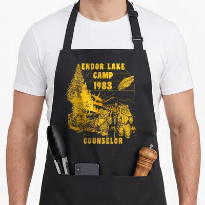 Endor camp counselor apron - Yellow retro print of two Ewoks, speeder bike, and Endor Lake Camp 1983 Counselor text — shown on a black cotton canvas apron against a white background