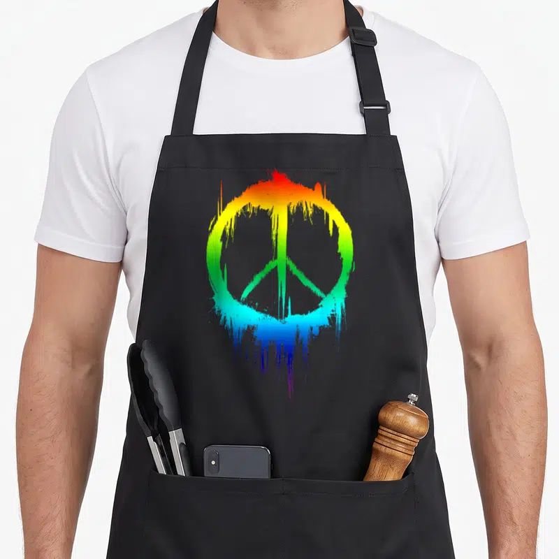 Peace paint apron - Rainbow peace symbol with red yellow green blue purple paint splatter and drip effect — shown on a black cotton canvas apron against a white background