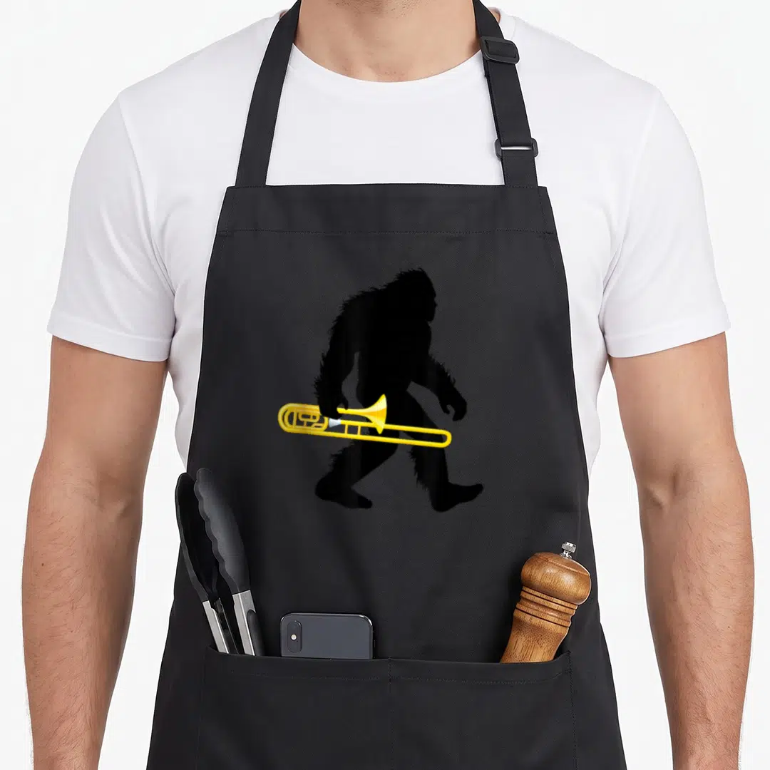 Trombone bigfoot apron - Black Bigfoot silhouette walking right holding yellow trombone cartoon — shown on a black cotton canvas apron against a white background