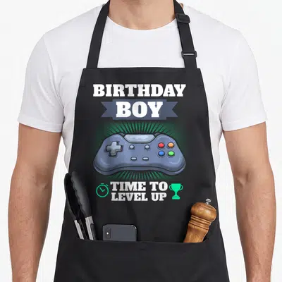 Theme video game apron - Birthday Boy Time to Level Up text with blue-grey game controller and mint icons on black bib — shown on a black cotton canvas apron against a white background