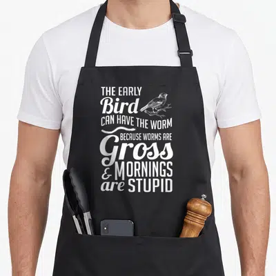 Early bird apron - White stacked text reading THE EARLY BIRD CAN HAVE THE WORM with bird and worm illustrations — shown on a black cotton canvas apron against a white background
