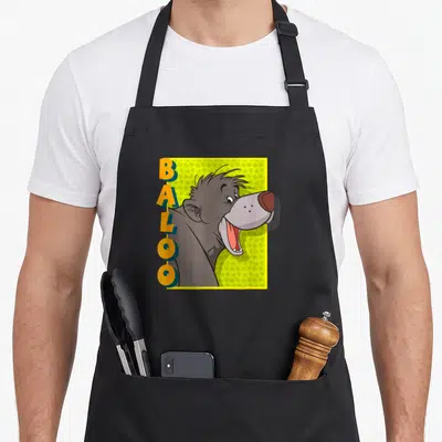 Jungle book apron - Vertical BALOO text beside grey cartoon bear head on yellow square with green leaf pattern — shown on a black cotton canvas apron against a white background