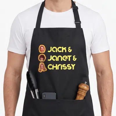 Jack face apron - Three stacked retro headshots with yellow JACK JANET CHRISSY text and orange outlines — shown on a black cotton canvas apron against a white background