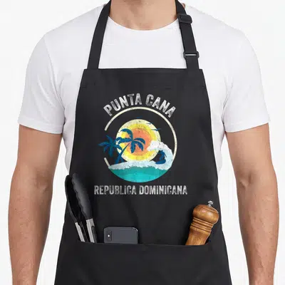 Republic punta cana apron - Retro circular beach graphic with surfer, sun, palms, and birds in white, blue, yellow — shown on a black cotton canvas apron against a white background