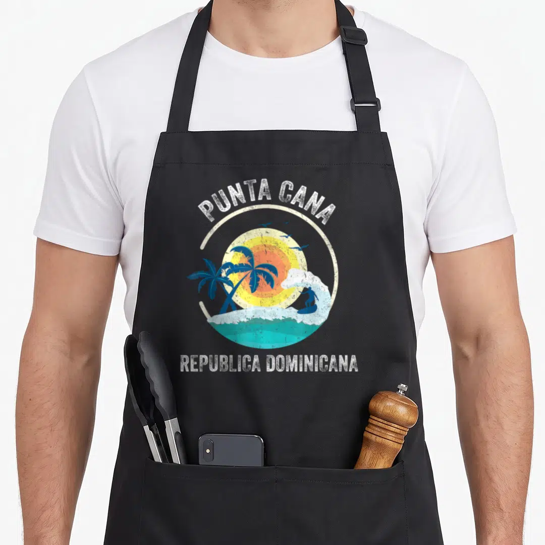 Republic punta cana apron - Retro circular beach graphic with surfer, sun, palms, and birds in white, blue, yellow — shown on a black cotton canvas apron against a white background