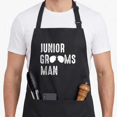 Groomsman junior apron - White stacked text JUNIOR GROOMSMAN MAN with sunglasses replacing the O letters in distressed — shown on a black cotton canvas apron against a white background