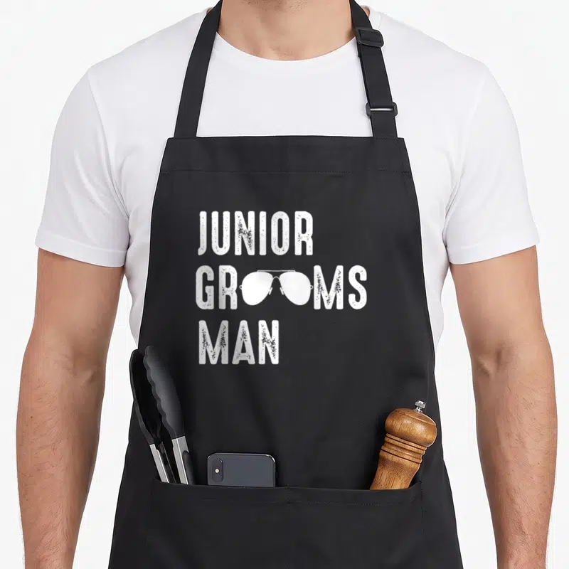 Groomsman junior apron - White stacked text JUNIOR GROOMSMAN MAN with sunglasses replacing the O letters in distressed — shown on a black cotton canvas apron against a white background