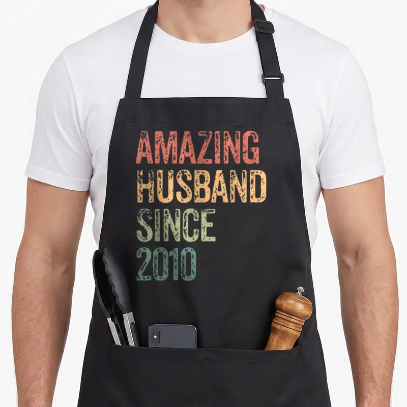 15th anniversary wedding apron - Stacked text Amazing Husband Since 2010 in orange, red, yellow, and green distressed — shown on a black cotton canvas apron against a white background