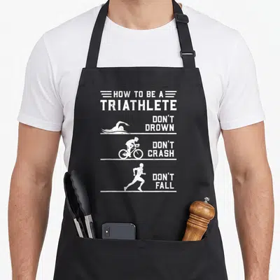 Triathlon triathlete apron - White text How To Be A Triathlete with swimmer cyclist runner silhouettes separated — shown on a black cotton canvas apron against a white background