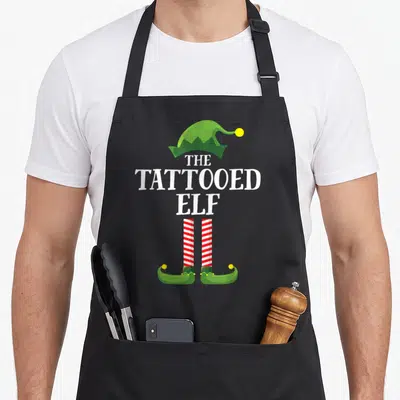 Tattooed elf apron - Centered white text THE TATTOOED ELF with green hat and striped legs below — shown on a black cotton canvas apron against a white background