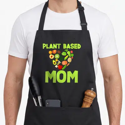 Mother plant based apron - Heart shape formed by fruits and vegetables with PLANT BASED MOM text in lime green — shown on a black cotton canvas apron against a white background
