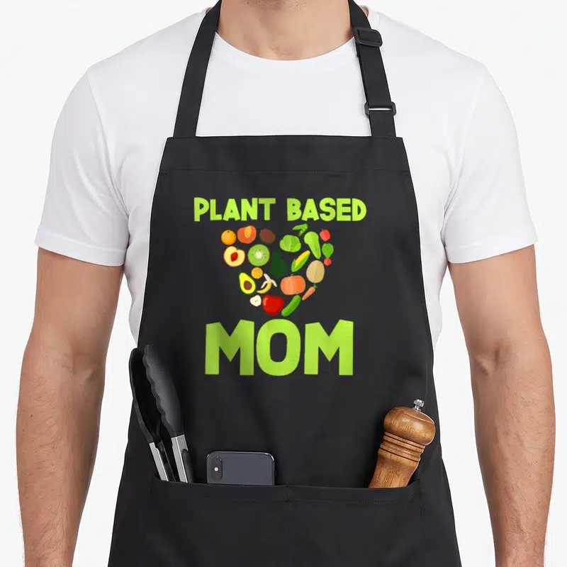 Mother plant based apron - Heart shape formed by fruits and vegetables with PLANT BASED MOM text in lime green — shown on a black cotton canvas apron against a white background