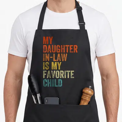 Law mother daughter apron - Stacked text MY DAUGHTER IN-LAW IS MY FAVORITE CHILD in red orange yellow beige and teal — shown on a black cotton canvas apron against a white background
