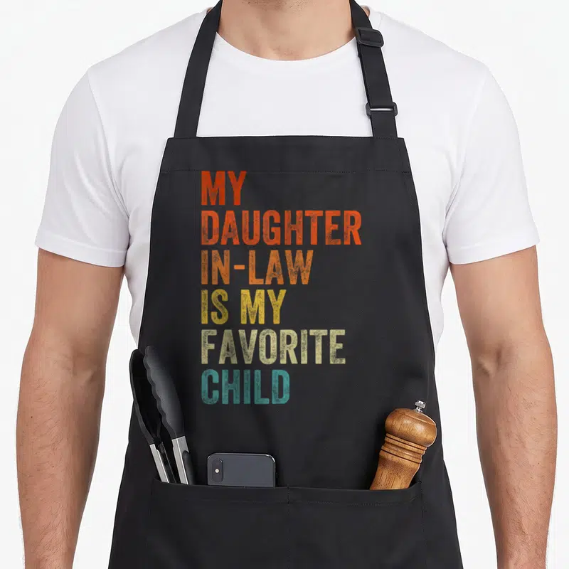 Law mother daughter apron - Stacked text MY DAUGHTER IN-LAW IS MY FAVORITE CHILD in red orange yellow beige and teal — shown on a black cotton canvas apron against a white background