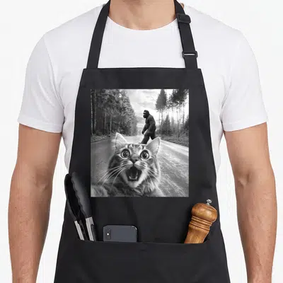 Foot funny cat apron - Surprised cat face close-up with Bigfoot walking on a forest road in black and white — shown on a black cotton canvas apron against a white background