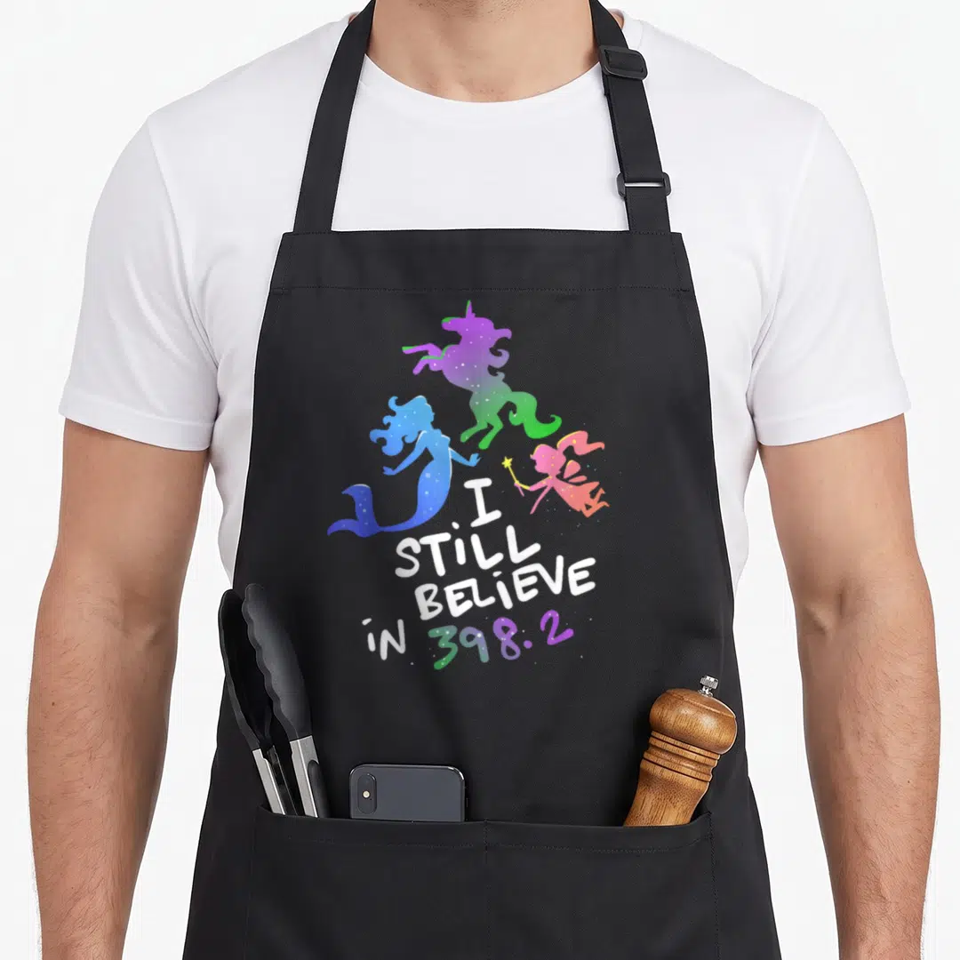 Believe distillery apron - I STILL BELIEVE in 398.2 text with purple unicorn, blue mermaid, pink fairy silhouettes — shown on a black cotton canvas apron against a white background