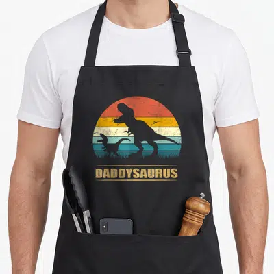 Daddysaurus daddy apron - Retro T-Rex silhouettes under striped sun with DADDYSAURUS text in grey yellow red teal — shown on a black cotton canvas apron against a white background