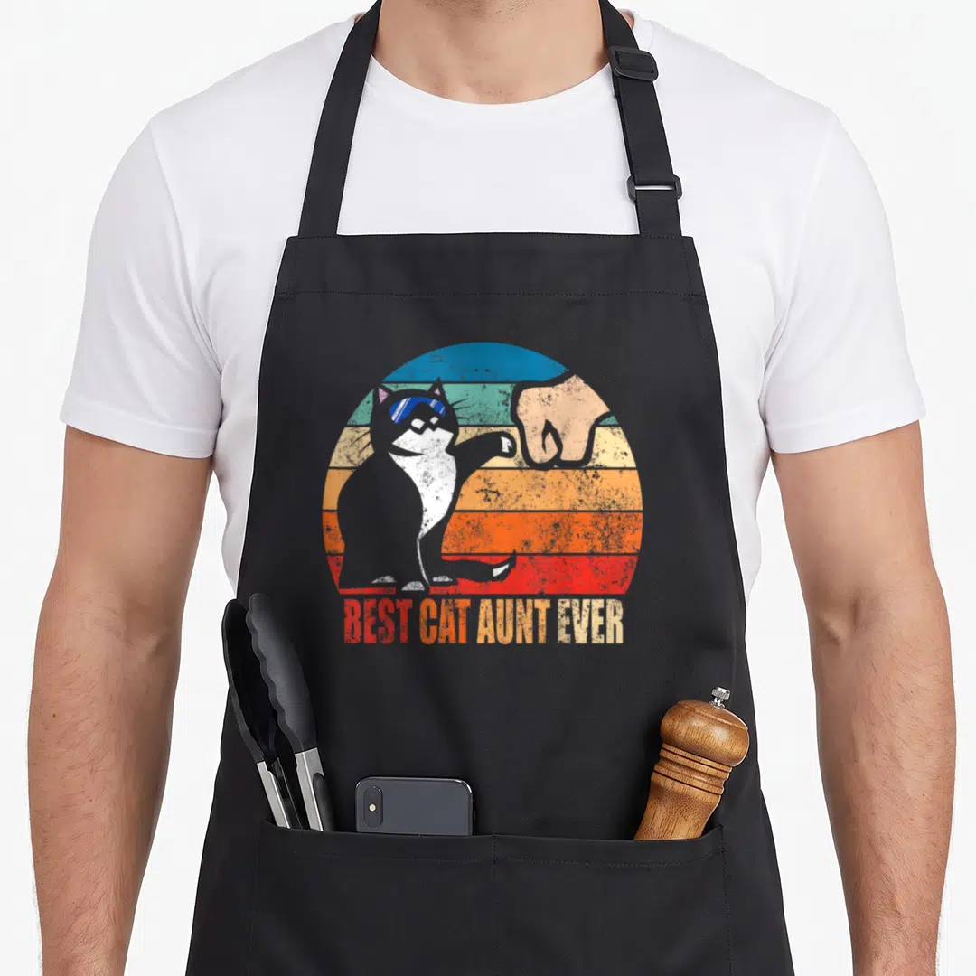 Aunt cat apron - Grey cat fist bumping human hand against blue orange red grey striped sun with text below — shown on a black cotton canvas apron against a white background