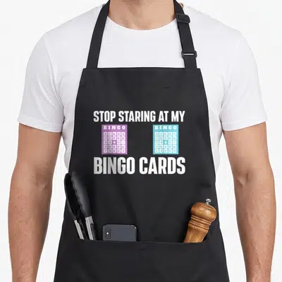 Stop bingo apron - Two bingo cards in purple and turquoise with white text reading stop staring at my bingo cards — shown on a black cotton canvas apron against a white background