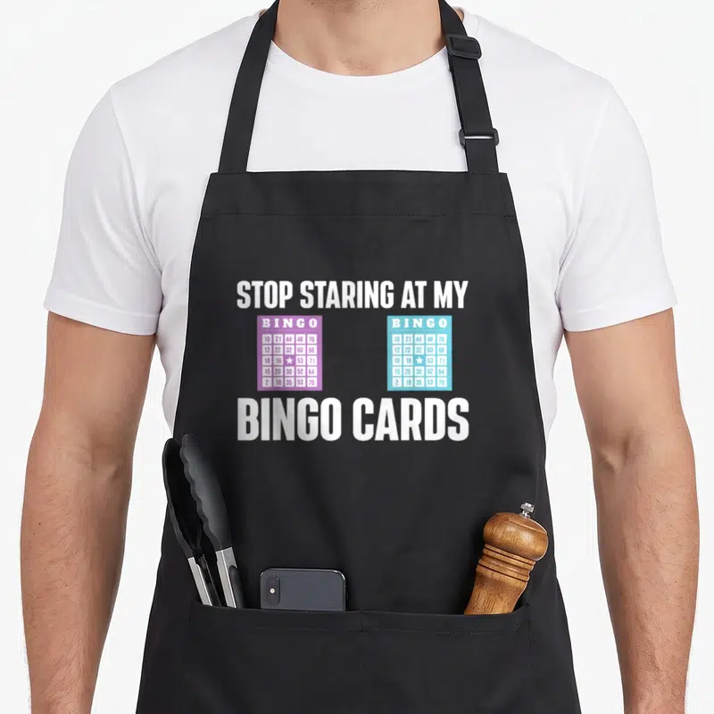 Stop bingo apron - Two bingo cards in purple and turquoise with white text reading stop staring at my bingo cards — shown on a black cotton canvas apron against a white background