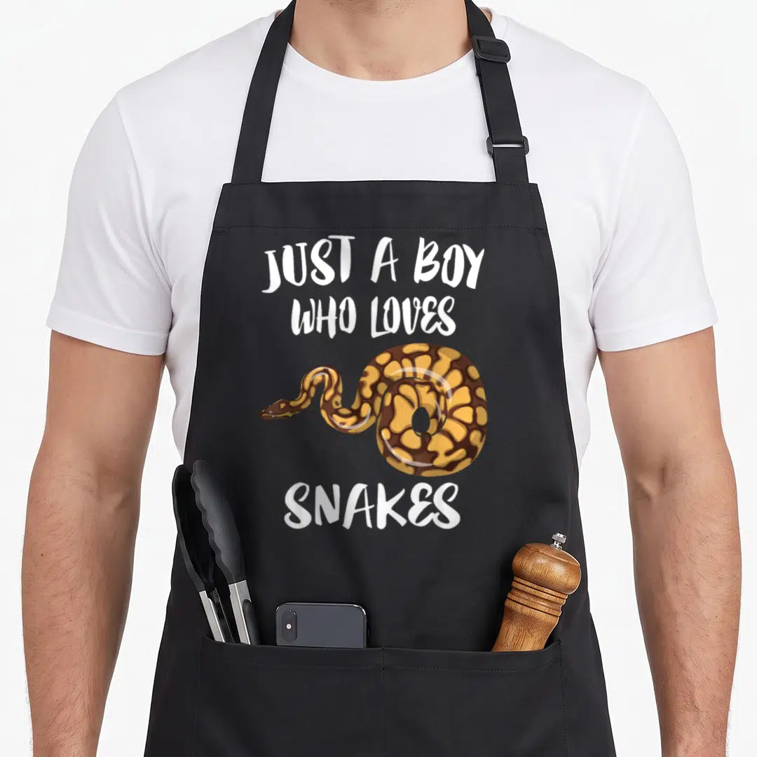 Snake animal apron - Cartoon brown and yellow snake with white text reading JUST A BOY WHO LOVES SNAKES — shown on a black cotton canvas apron against a white background