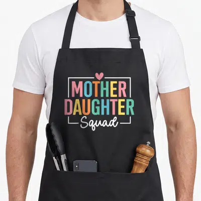 Funny daughter my mother's apron - MOTHER DAUGHTER Squad text with pink heart and white frame in rainbow colors — shown on a black cotton canvas apron against a white background