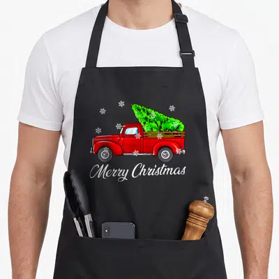 Farmer christmas tree apron - Red truck carrying green Christmas tree with white Merry Christmas script and snowflakes — shown on a black cotton canvas apron against a white background