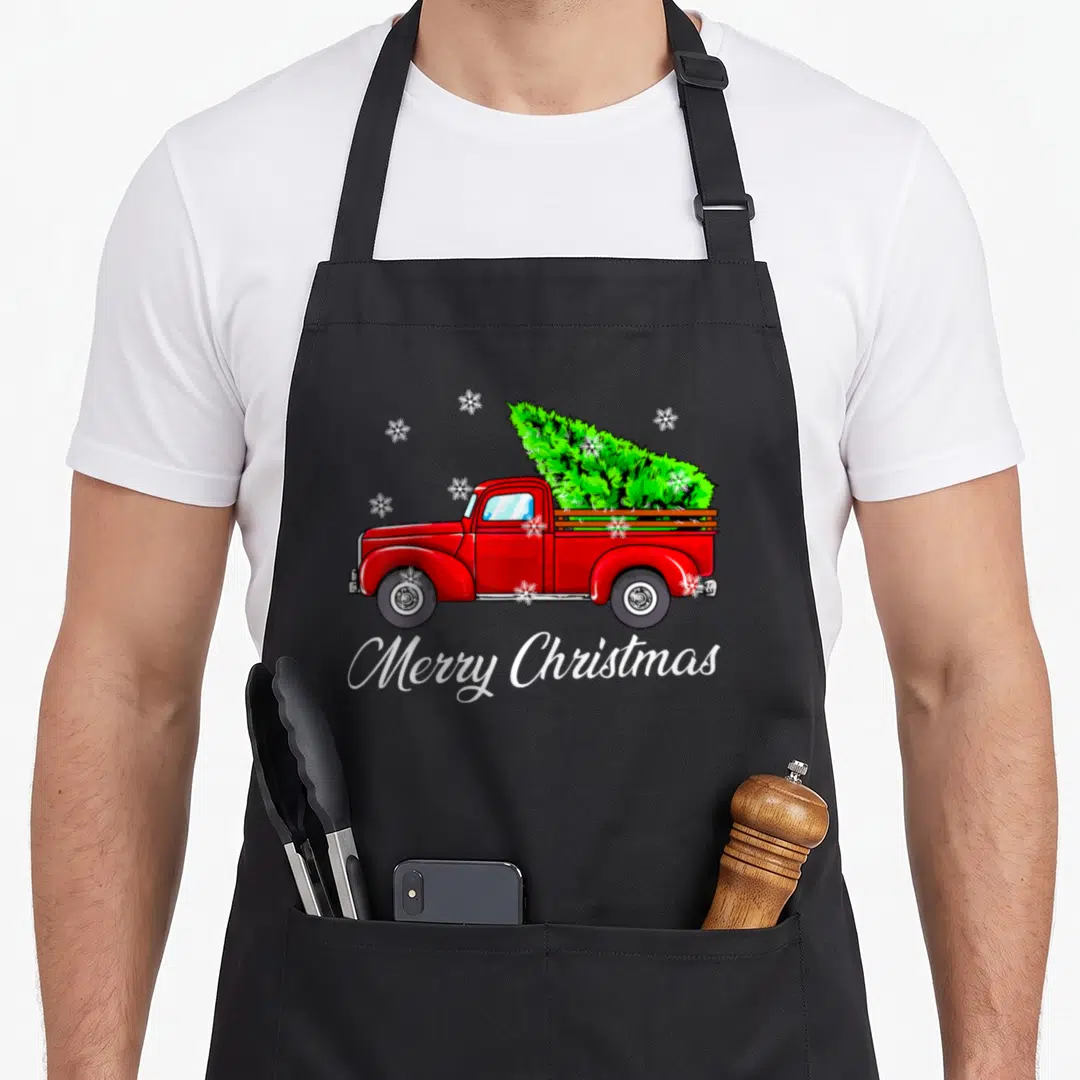 Farmer christmas tree apron - Red truck carrying green Christmas tree with white Merry Christmas script and snowflakes — shown on a black cotton canvas apron against a white background