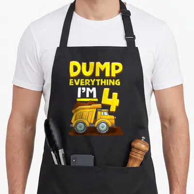 4th dump truck apron - DUMP EVERYTHING I'M 4 text above yellow dump truck on brown dirt pile — shown on a black cotton canvas apron against a white background