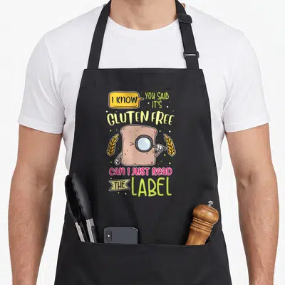 Gluten lifestyle apron - Cartoon bread slice with magnifying glass and text asking to read the label — shown on a black cotton canvas apron against a white background