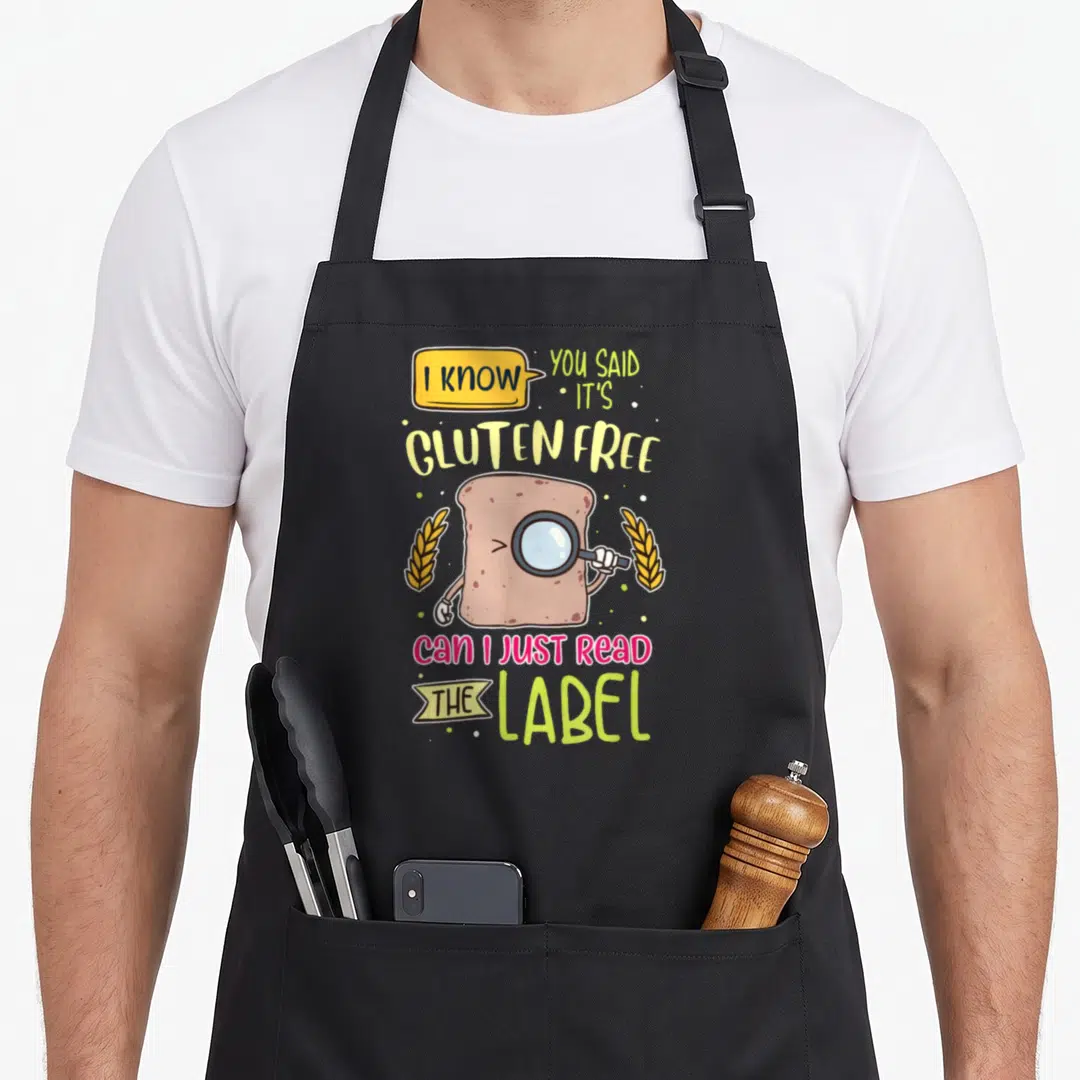 Gluten lifestyle apron - Cartoon bread slice with magnifying glass and text asking to read the label — shown on a black cotton canvas apron against a white background