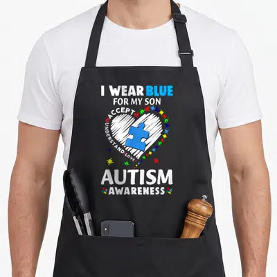 Blue nurse dad apron - I WEAR BLUE FOR MY SON text around white heart with blue puzzle piece and colorful fragments — shown on a black cotton canvas apron against a white background