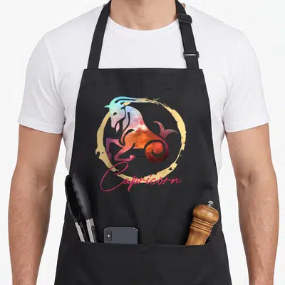 Astrology capricorn zodiac aprons - Galaxy sea-goat inside gold circle with pink Capricorn script text — shown on a black cotton canvas apron against a white background