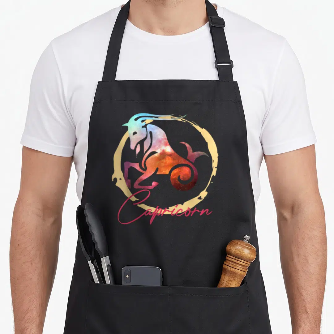 Astrology capricorn zodiac aprons - Galaxy sea-goat inside gold circle with pink Capricorn script text — shown on a black cotton canvas apron against a white background