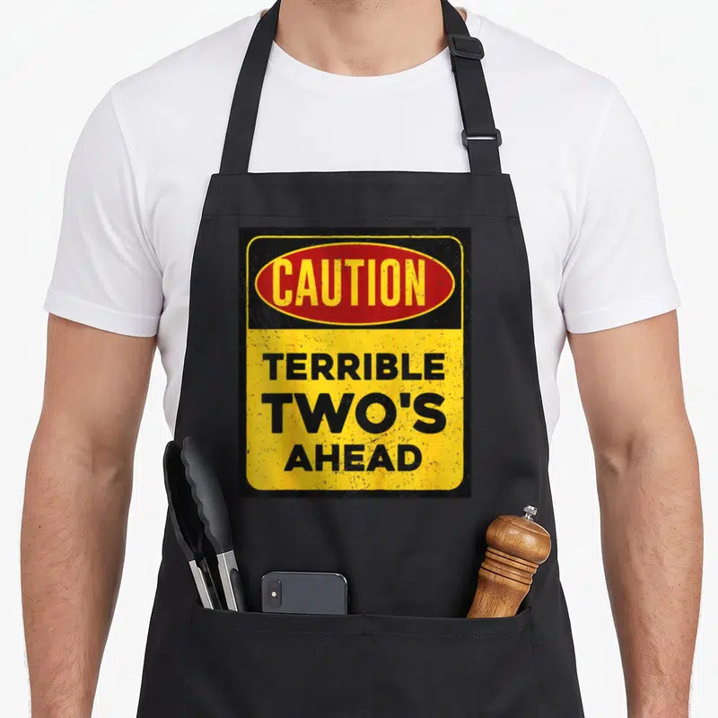 Caution construction apron - Road sign graphic with red oval CAUTION and stacked black text TERRIBLE TWO'S AHEAD — shown on a black cotton canvas apron against a white background