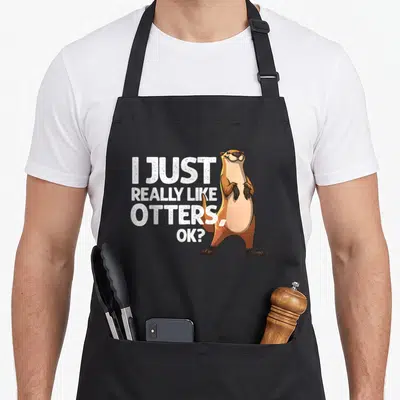 Badger animal apron - I Just Really Like Otters, Ok? text next to a standing cartoon otter in tan and brown — shown on a black cotton canvas apron against a white background