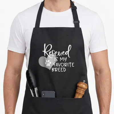 Rescued pet owner apron - White text Rescued is my favorite breed next to a heart with a paw print inside — shown on a black cotton canvas apron against a white background