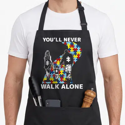 Youll walking apron - You'll Never Walk Alone text with family silhouettes and multi-colored puzzle piece ribbon — shown on a black cotton canvas apron against a white background