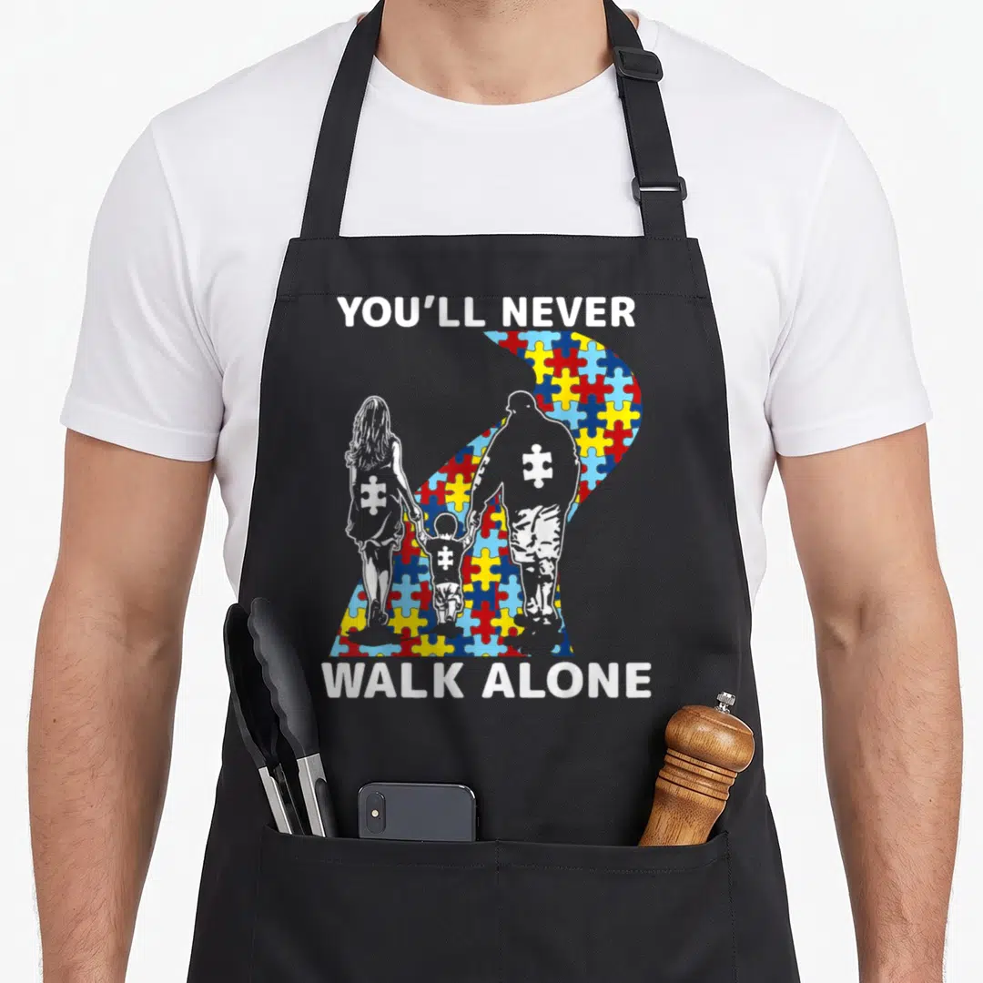 Youll walking apron - You'll Never Walk Alone text with family silhouettes and multi-colored puzzle piece ribbon — shown on a black cotton canvas apron against a white background